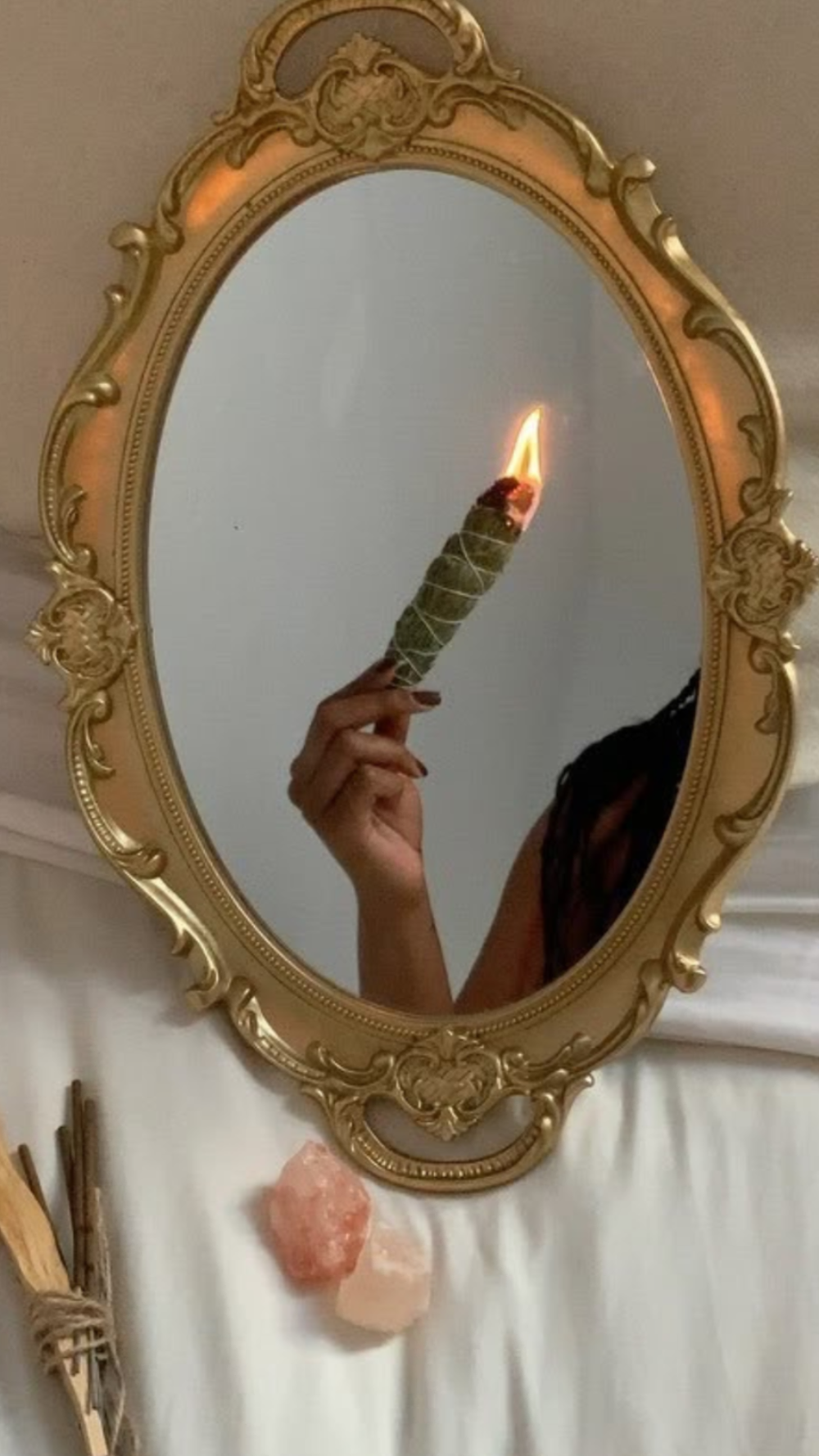 An ornate gold oval mirror reflecting a hand holding a lit sage bundle candle, set against a white draped fabric background with pink crystal stones and incense sticks below.