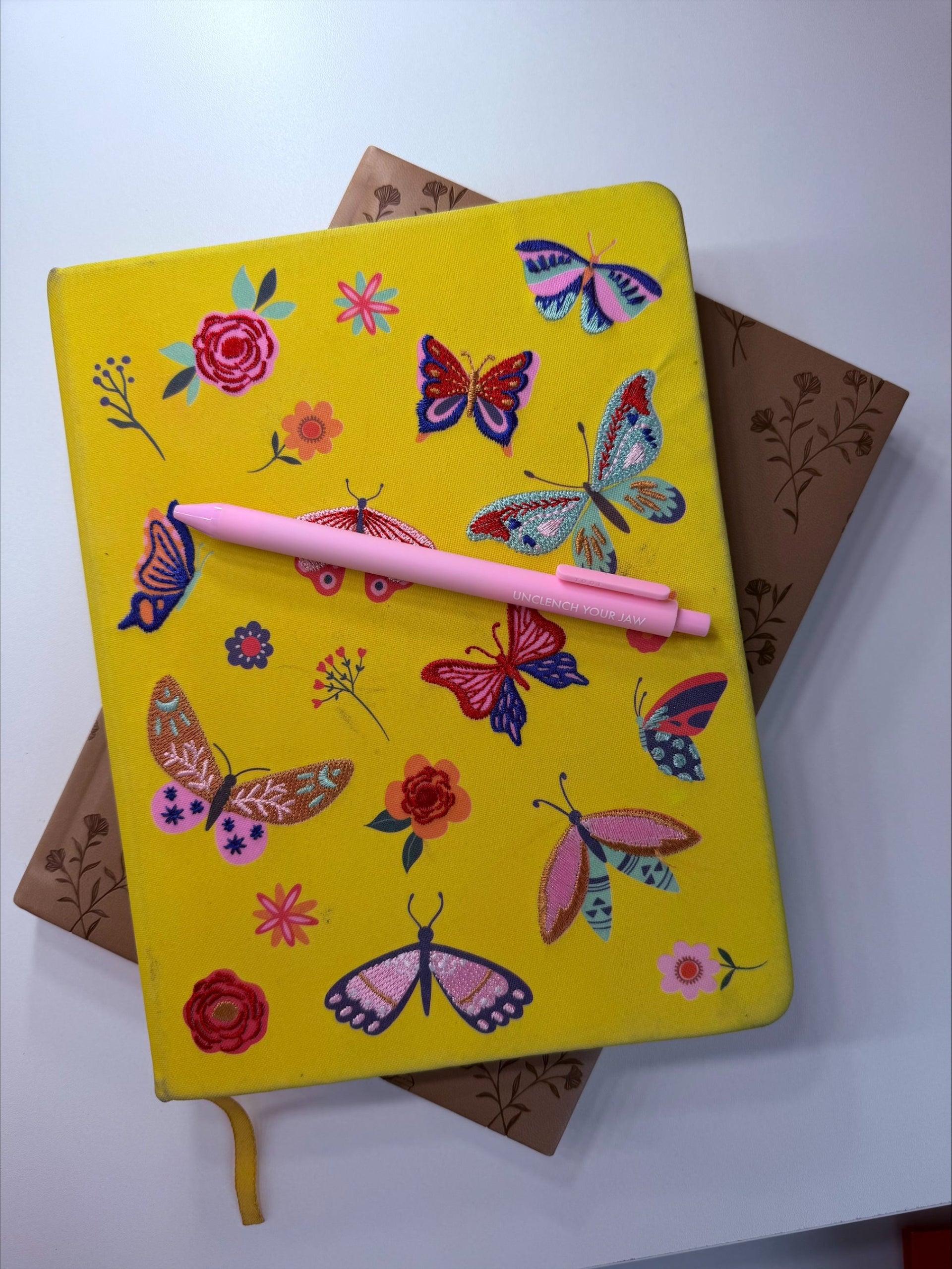 A bright yellow hardcover notebook decorated with colorful butterflies and flowers, resting on top of brown floral-patterned books with a pink pen placed across the cover.